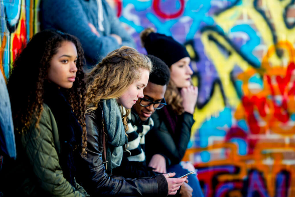 A group of teenagers are sitting in front of a wall covered in graffiti. They are wearing stylish clothes. A boy and girl are looking at a smartphone screen together.